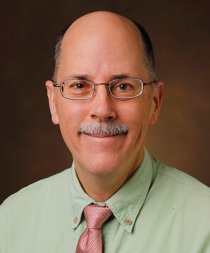 Headshot of Chuck Sanders, who is wearing a light red/pink tie with blue highlights and a green collared shirt.